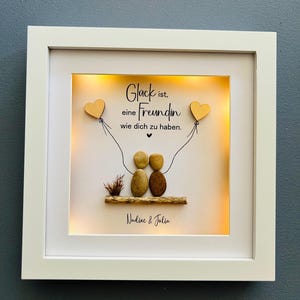 May include: A white framed picture with a light inside. The picture has a quote that reads "Gl&uuml;ck ist, eine Freundin wie dich zu haben."  The quote is written in black lettering on a white background. There are two small wooden hearts with strings attached to them. The hearts are hanging from the top of the picture. There are two small brown stones that look like people sitting on a small piece of wood. The stones are holding hands. The picture is signed "Nadine & Julia".
