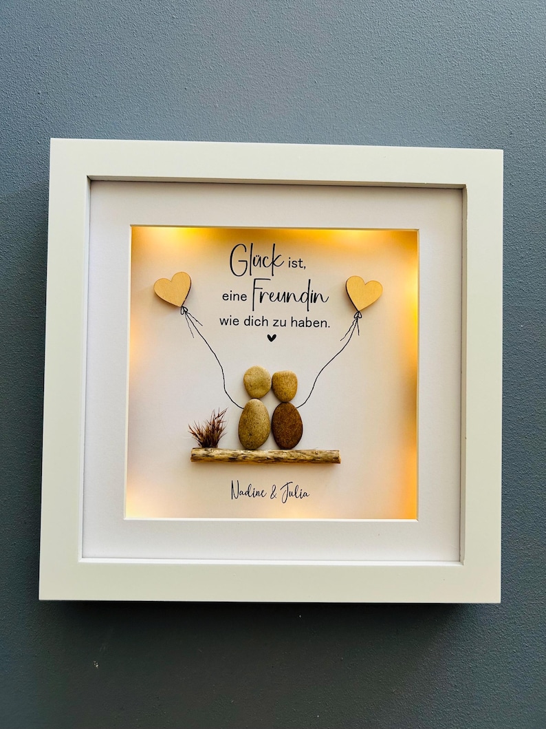 May include: A white framed picture with a light inside. The picture has a quote that reads "Gl&uuml;ck ist, eine Freundin wie dich zu haben."  The quote is written in black lettering on a white background. There are two small wooden hearts with strings attached to them. The hearts are hanging from the top of the picture. There are two small brown stones that look like people sitting on a small piece of wood. The stones are holding hands. The picture is signed "Nadine & Julia".