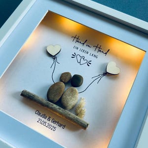 May include: A framed artwork with a white border. The artwork features a couple depicted with stones, holding wooden heart balloons. The text "Hand in Hand EIN LEBEN LANG" is written above the couple, with the names Claudia & Gerhard and the date 21.05.2025 below.