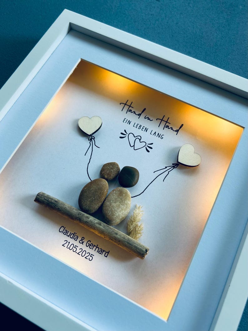 May include: A framed artwork with a white border. The artwork features a couple depicted with stones, holding wooden heart balloons. The text "Hand in Hand EIN LEBEN LANG" is written above the couple, with the names Claudia & Gerhard and the date 21.05.2025 below.