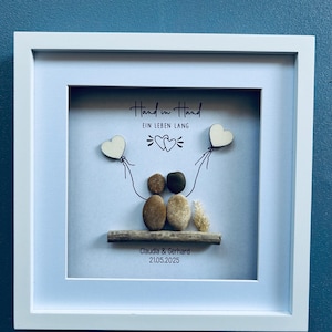 May include: Framed artwork with the text "Hand in Hand" and "Ein Leben Lang". The artwork features a couple made of stones, holding wooden heart balloons. The names "Claudia & Gerhard" and the date "21.05.2025" are also included.