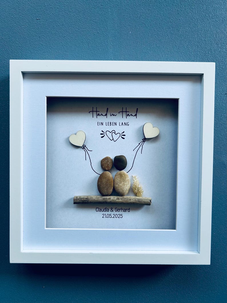 May include: Framed artwork with the text "Hand in Hand" and "Ein Leben Lang". The artwork features a couple made of stones, holding wooden heart balloons. The names "Claudia & Gerhard" and the date "21.05.2025" are also included.
