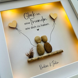 May include: A white frame with a light inside, displaying a hand-crafted artwork made of stones and driftwood. The artwork depicts two figures holding hands, with two wooden hearts attached to strings above them. The text "Gl&uuml;ck ist eine Freundin wie dich zu haben." is written in black lettering on a white background. The names "Nadine & Julia" are written below the artwork.