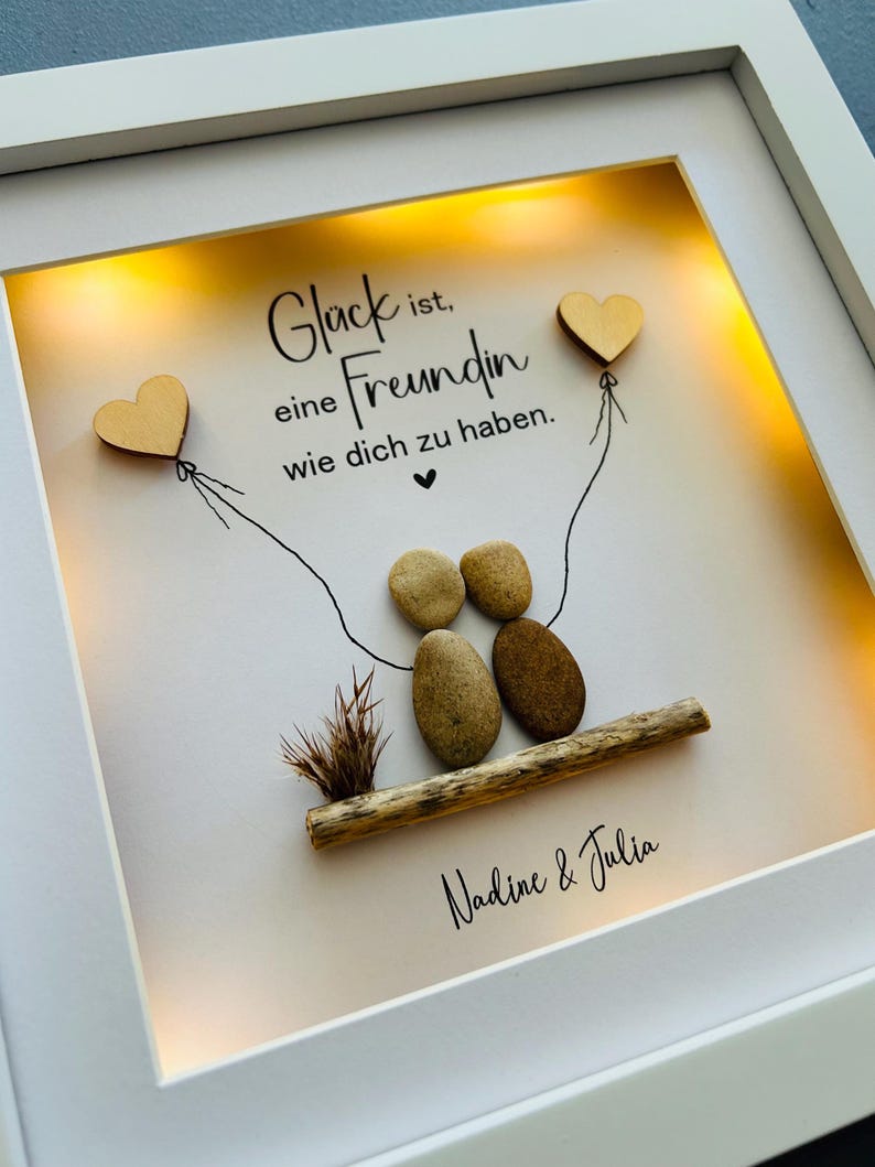 May include: A white frame with a light inside, displaying a hand-crafted artwork made of stones and driftwood. The artwork depicts two figures holding hands, with two wooden hearts attached to strings above them. The text "Gl&uuml;ck ist eine Freundin wie dich zu haben." is written in black lettering on a white background. The names "Nadine & Julia" are written below the artwork.