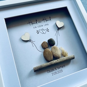 May include: A framed artwork with a white border. The artwork features a pebble art couple holding wooden heart balloons. The phrase "Hand in Hand" and "EIN LEBEN LANG" are written above the couple. The names "Claudia & Gerhard" and the date "21.05.2025" are below.