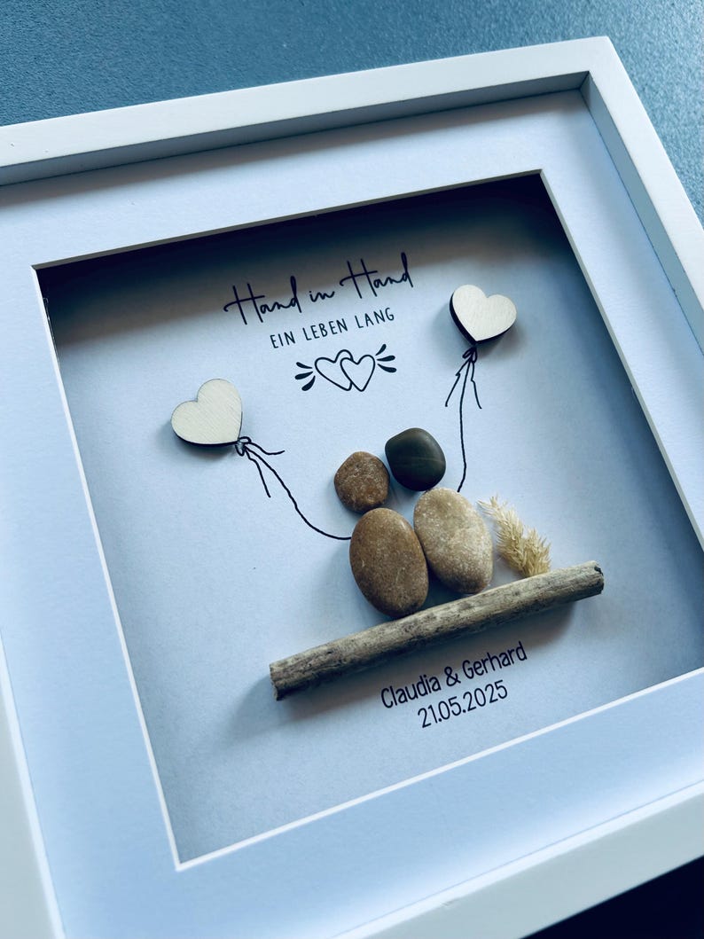 May include: A framed artwork with a white border. The artwork features a pebble art couple holding wooden heart balloons. The phrase "Hand in Hand" and "EIN LEBEN LANG" are written above the couple. The names "Claudia & Gerhard" and the date "21.05.2025" are below.
