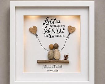May include: A framed artwork with a white border features a heartwarming design. The artwork includes the text "Liebe ist, wenn aus dem Ich & Du ein Wir entsteht." Two wooden heart-shaped balloons are connected to a couple made of stones. The names "Stefanie & Michael" and the date "05.04.2026" are also included.