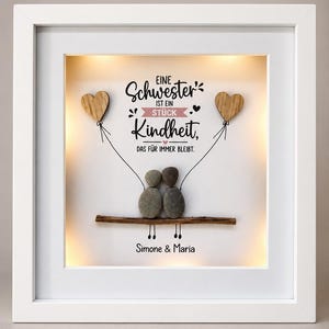 May include: A white square shadow box frame containing a pebble art piece of two figures sitting on a twig, holding strings attached to two wooden hearts. The text reads: EINE Schwester IST EIN STÜCK Kindheit, DAS FÜR IMMER BLEIBT. Simone & Maria.