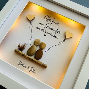 May include: A white frame with a light inside, displaying a hand-drawn illustration of two hearts connected by a string. The text "Gl&uuml;ck ist, eine Freundin wie dich zu haben." is written in black ink. The names "Nadine & Julia" are written below the illustration.