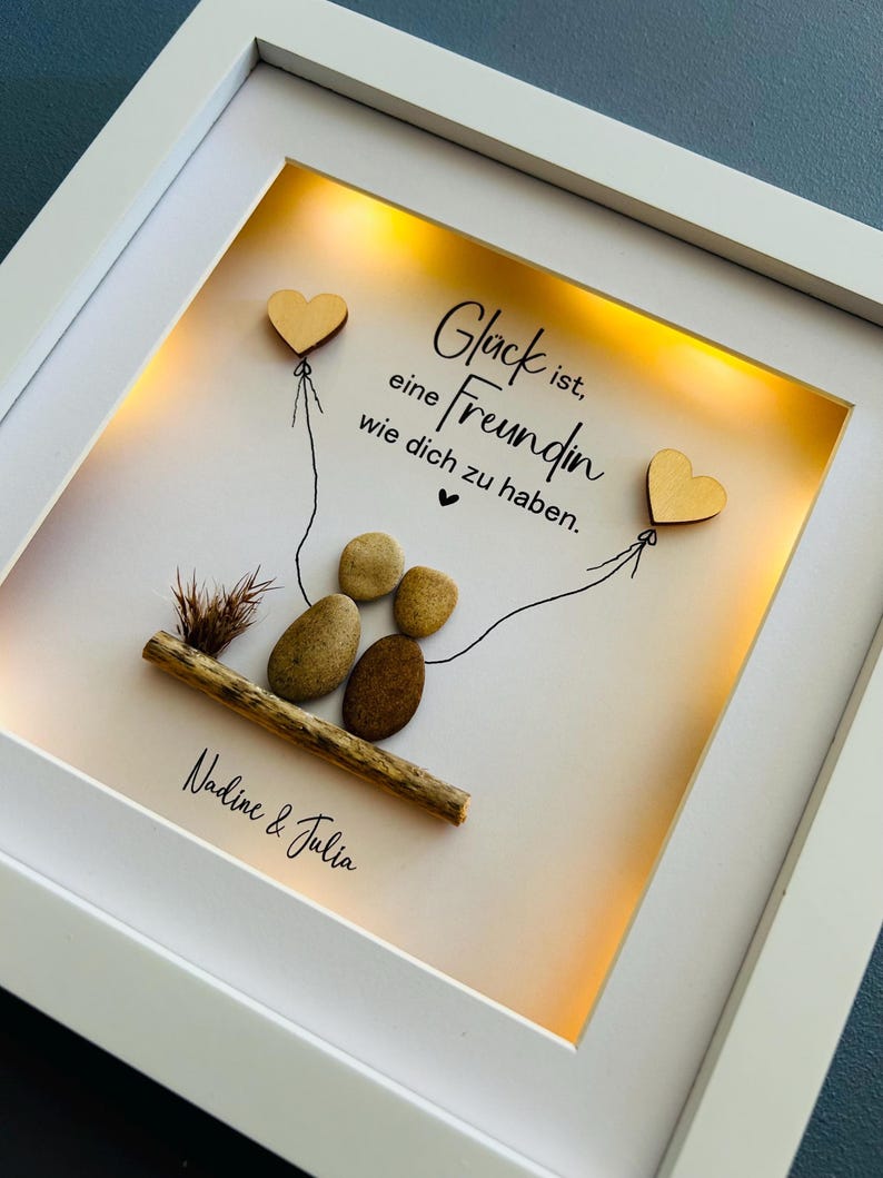 May include: A white frame with a light inside, displaying a hand-drawn illustration of two hearts connected by a string. The text "Gl&uuml;ck ist, eine Freundin wie dich zu haben." is written in black ink. The names "Nadine & Julia" are written below the illustration.