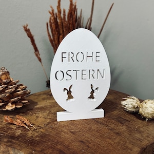 Könnte beinhalten: Ein weißes, hölzernes Osterei-Schild mit dem Text "Frohe Ostern" und zwei Hasensilhouetten.