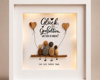 May include: A square white framed wall art piece featuring a pebble art family of four sitting on a wooden branch. The text reads "Glück ist... Großeltern wie euch zu haben!" with the names "Luis Lutz Kathrin Noah" below. Two wooden heart balloons are attached by thin lines.