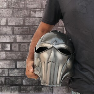Op de afbeelding: Een persoon die een zwarte metalen helm vasthoudt met een gezichtsmasker en een vizier. De helm heeft een getextureerd oppervlak en een puntige bovenkant.