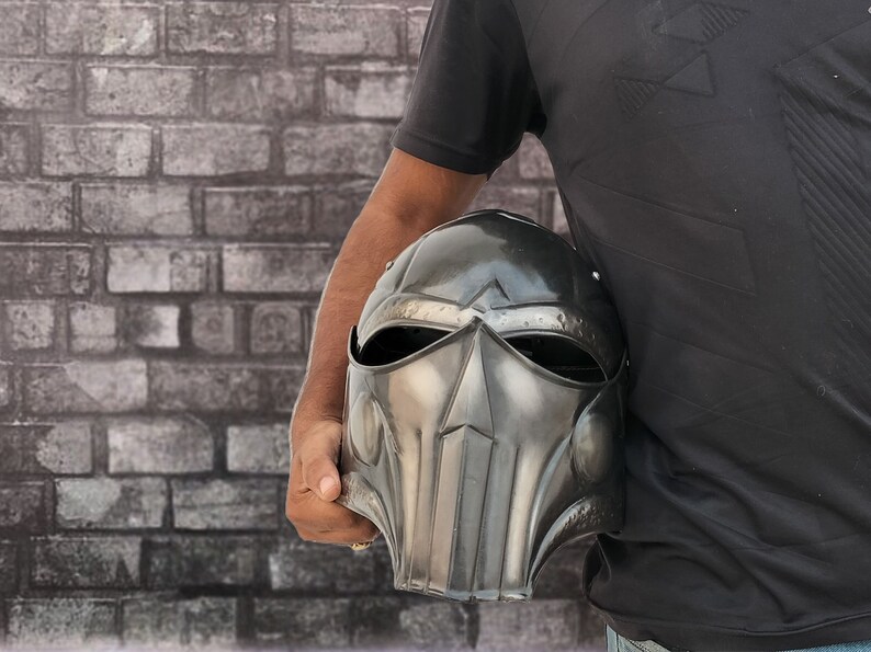 Op de afbeelding: Een persoon die een zwarte metalen helm vasthoudt met een gezichtsmasker en een vizier. De helm heeft een getextureerd oppervlak en een puntige bovenkant.