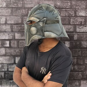 Op de afbeelding: Een persoon draagt een zwart T-shirt met een wit "NY"-logo en een donkergrijze helm met een vizier. De helm heeft een getextureerd oppervlak en lijkt van metaal te zijn.