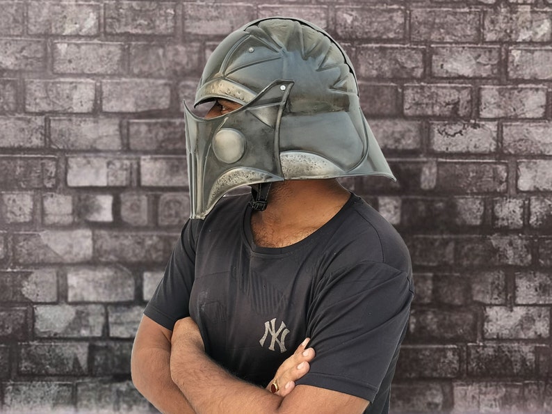 Op de afbeelding: Een persoon draagt een zwart T-shirt met een wit "NY"-logo en een donkergrijze helm met een vizier. De helm heeft een getextureerd oppervlak en lijkt van metaal te zijn.