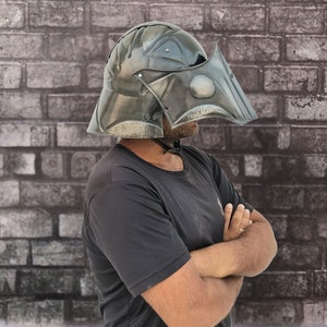 Op de afbeelding: Een persoon draagt ​​een donkergrijze helm met een vizier. De helm heeft een getextureerd oppervlak en een puntig ontwerp. De persoon staat voor een bakstenen muur.