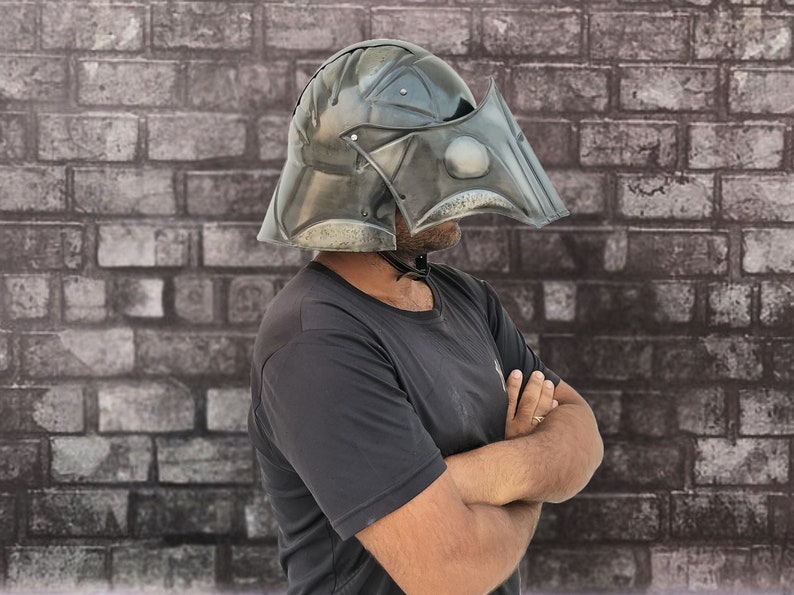 Op de afbeelding: Een persoon draagt ​​een donkergrijze helm met een vizier. De helm heeft een getextureerd oppervlak en een puntig ontwerp. De persoon staat voor een bakstenen muur.