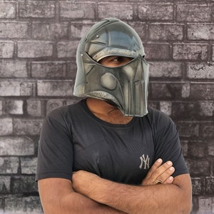 Op de afbeelding: Een persoon draagt ​​een donkergrijze helm met een gezichtsbeschermer. De helm heeft een getextureerd oppervlak en een ontwerp aan de voorkant. De persoon draagt ​​een zwart T-shirt met een wit logo.