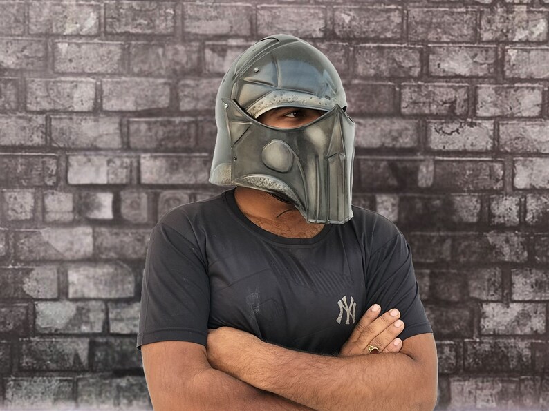 Op de afbeelding: Een persoon draagt ​​een donkergrijze helm met een gezichtsbeschermer. De helm heeft een getextureerd oppervlak en een ontwerp aan de voorkant. De persoon draagt ​​een zwart T-shirt met een wit logo.
