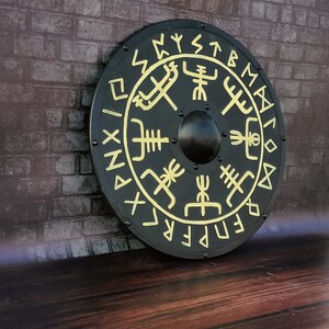 Könnte beinhalten: Ein runder schwarzer Schild mit goldenen Runen und Symbolen. Der Schild hat eine erhabene Mitte und ist mit verschiedenen nordischen Symbolen verziert.