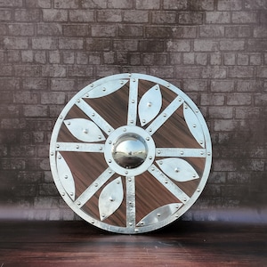 Könnte beinhalten: Ein runder Holzschild mit einem silbernen Metallrahmen und dekorativen Akzenten. Der Schild hat einen zentralen silbernen Metallboss.