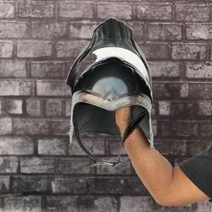 Op de afbeelding: Een zwarte helm met een puntige bovenkant en een metalen band rond de onderkant. De helm wordt in de hand van een persoon gehouden.
