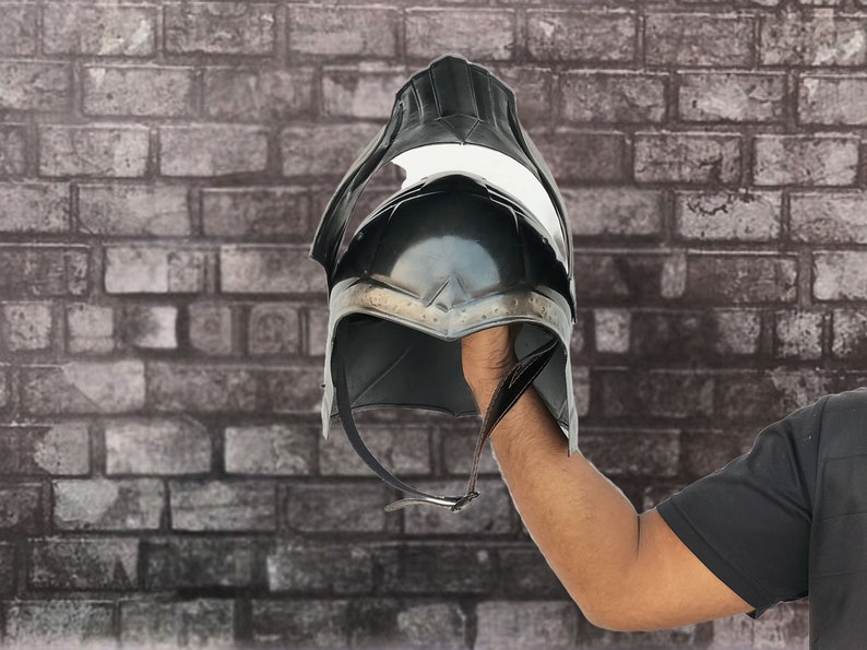 Op de afbeelding: Een zwarte helm met een puntige bovenkant en een metalen band rond de onderkant. De helm wordt in de hand van een persoon gehouden.