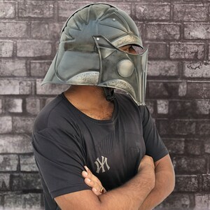 Op de afbeelding: Een persoon draagt een zwart T-shirt met een wit logo en een donkergrijze helm met een vizier. De helm heeft een getextureerd oppervlak en lijkt van metaal te zijn.