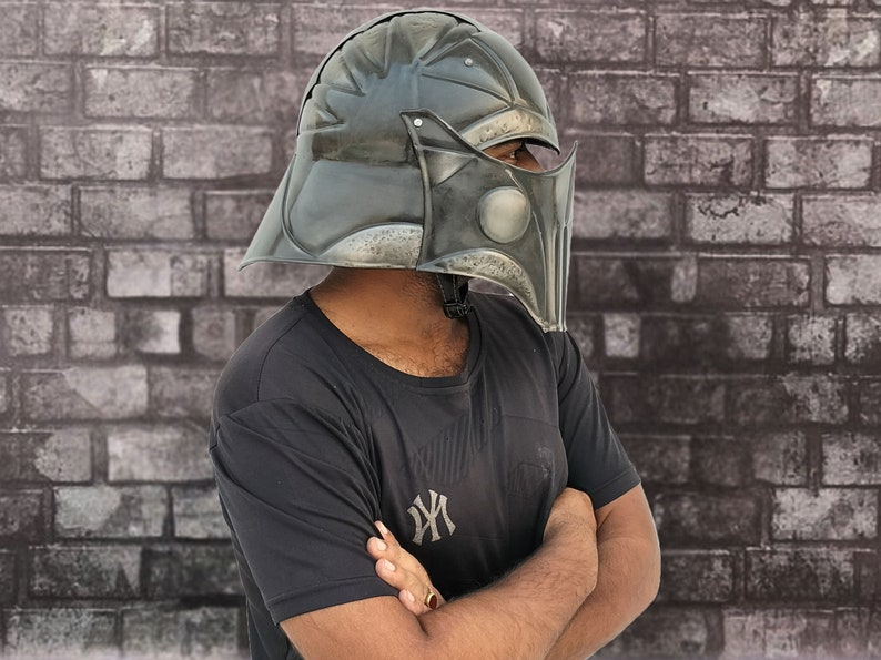 Op de afbeelding: Een persoon draagt een zwart T-shirt met een wit logo en een donkergrijze helm met een vizier. De helm heeft een getextureerd oppervlak en lijkt van metaal te zijn.