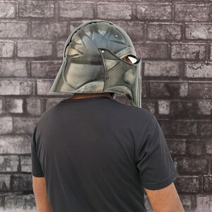 Op de afbeelding: Een persoon draagt ​​een donkergrijze helm met een vizier. De helm heeft een getextureerd oppervlak en lijkt van metaal te zijn. De persoon staat voor een bakstenen muur.