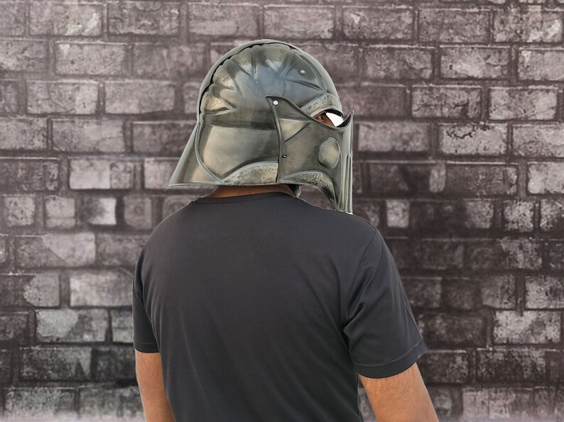 Op de afbeelding: Een persoon draagt ​​een donkergrijze helm met een vizier. De helm heeft een getextureerd oppervlak en lijkt van metaal te zijn. De persoon staat voor een bakstenen muur.