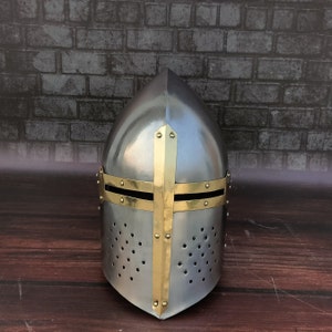 May include: A silver helmet with a brass cross design. The helmet has a pointed top and a face shield with small holes.
