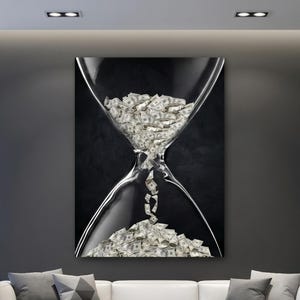 May include: A large canvas art print shows an hourglass filled with money, representing time and wealth. The artwork has a black background and is displayed above a white sofa in a modern living room setting.