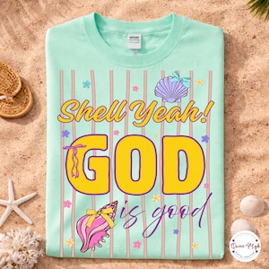 Op de afbeelding: Een lichtgroen t-shirt met de woorden "Shell Yeah! GOD is good" in geel en paars. Het shirt heeft schelpgraphics en verticale strepen in roze en wit. Het shirt wordt gepresenteerd op een zanderige ondergrond met strandaccessoires.