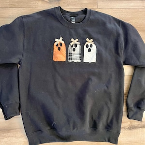 May include: A black crewneck sweatshirt featuring three ghost-shaped fabric appliqués on the chest. The ghosts are orange, black and white plaid, and white, each topped with a small tan bow and featuring black stitched eyes and open mouths.