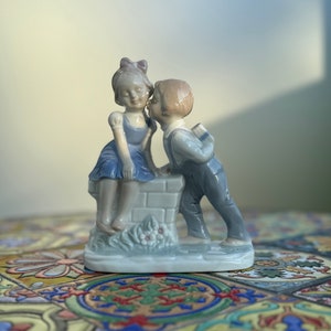 Peut inclure: Une figurine en porcelaine d'un garçon et d'une fille qui s'embrassent. Le garçon porte une salopette bleue et la fille une robe bleue. La figurine est assise sur un petit mur de briques.