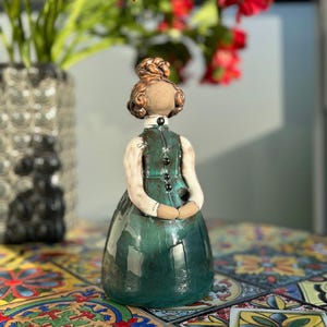 Pode incluir: Estatueta de cerâmica de uma mulher em um vestido longo verde-azulado, gola alta e botões pretos. Seus braços estão cruzados e seu cabelo está preso em um coque. A estatueta está em uma superfície de azulejos coloridos, com flores vermelhas ao fundo.