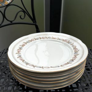 Può includere: Sei piatti da cena in ceramica bianca impilati con un bordo dorato e un motivo floreale in tonalità rosa, blu e marrone.
