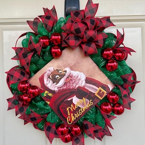 Puede incluir: Una corona de Navidad con una cinta de cuadros rojos y negros, malla verde y adornos rojos. Una imagen vintage de un Papá Noel negro está en el centro con el texto "Feliz Navidad".
