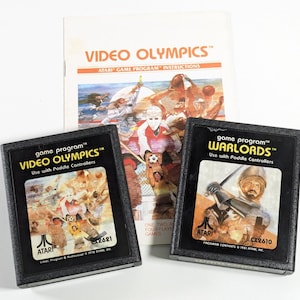 May include: Two Atari video game cartridges, one for "Video Olympics" and one for "Warlords", both with black cases and colorful artwork. The cartridges are accompanied by a white instruction booklet for "Video Olympics".
