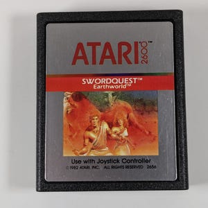 May include: A black cartridge for the Atari 2600 video game console. The cartridge has a silver label with the text "ATARI 2600", "SWORDQUEST™", and "Earthworld™". The label also includes a picture of two people in a fantasy setting.