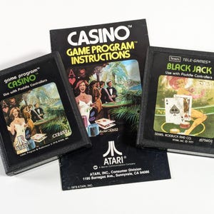 May include: Three Atari video game cartridges, two are for the game "Casino" and one is for "Black Jack". The cartridges are black with white text and images. The cartridges are on top of a black and white instruction booklet for the game "Casino".