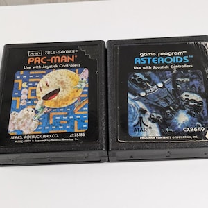 May include: Two black video game cartridges. One features Pac-Man with a yellow character and blue maze. The other shows a space scene with the word "Asteroids". Both are labeled to use with joystick controllers.
