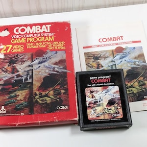 May include: A red box with a picture of a fighter jet and tanks on the front. The box is labeled "COMBAT Video Computer System Game Program" and "27 Video Games". The box also has the Atari logo and the number CX2601. There is a black cartridge with the text "COMBAT Game Program" and "CX2601" on it.
