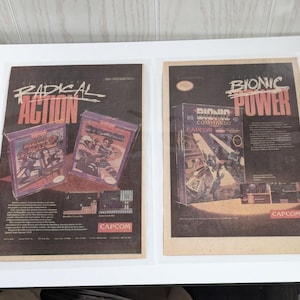 May include: Two vintage video game advertisements. The left ad features the game "Radical Action" with a red, white, and blue color scheme. The right ad features the game "Bionic Commando" with a red, white, and black color scheme. Both ads feature the Capcom logo.