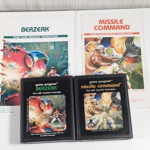 May include: Two Atari game cartridges, one for the game "Berzerk" and one for the game "Missile Command". Both cartridges are black with white text and graphics. The cartridges are on a white surface.