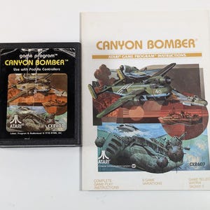 May include: A black Atari game cartridge with the text "game program" "CANYON BOMBER" and "Use with Paddle Controllers". The cartridge is next to a folded instruction booklet with the text "CANYON BOMBER" and "ATARI GAME PROGRAM INSTRUCTIONS".