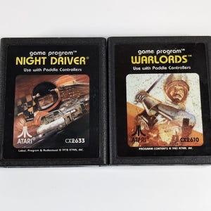 May include: Two vintage Atari game cartridges. The "NIGHT DRIVER" cartridge features a race car graphic, while the "WARLORDS" cartridge displays a medieval warrior. Both are black with white and orange text. The cartridges are labeled "Use with Paddle Controllers."