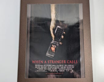May include: Movie poster for the film "When a Stranger Calls" featuring a hand holding a cell phone with a woman's face on the screen. The poster is in a brown frame.